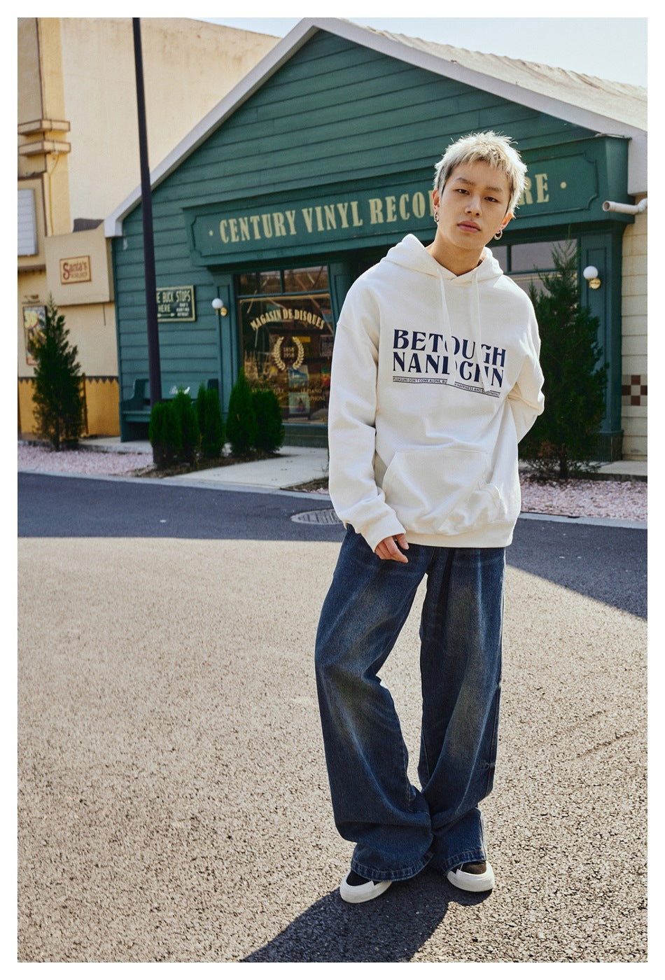 Model wearing a white pullover hoodie with a large dark chest graphic and a front pouch, styled with wide-leg blue jeans.