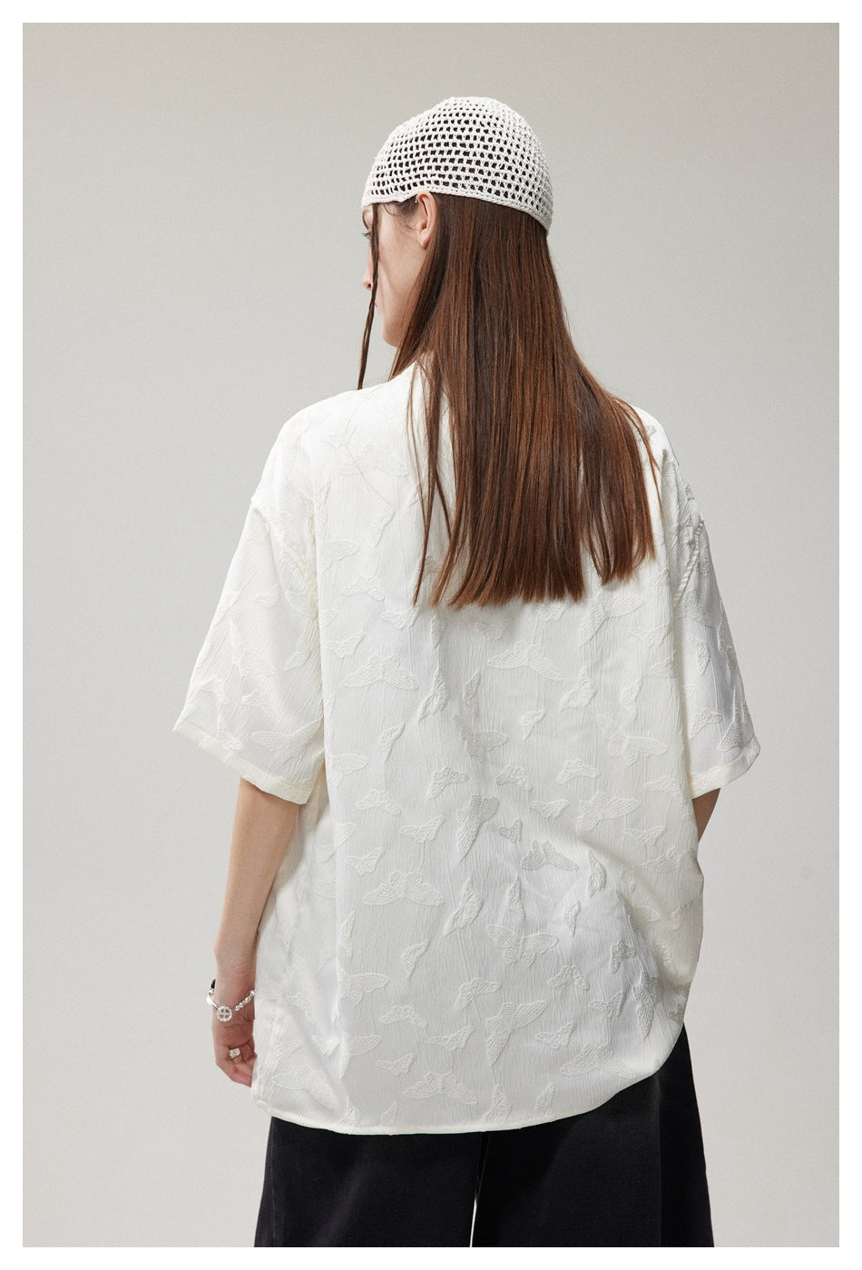 Back view of a model wearing an oversized white textured short-sleeve shirt with raised butterfly-like motifs.