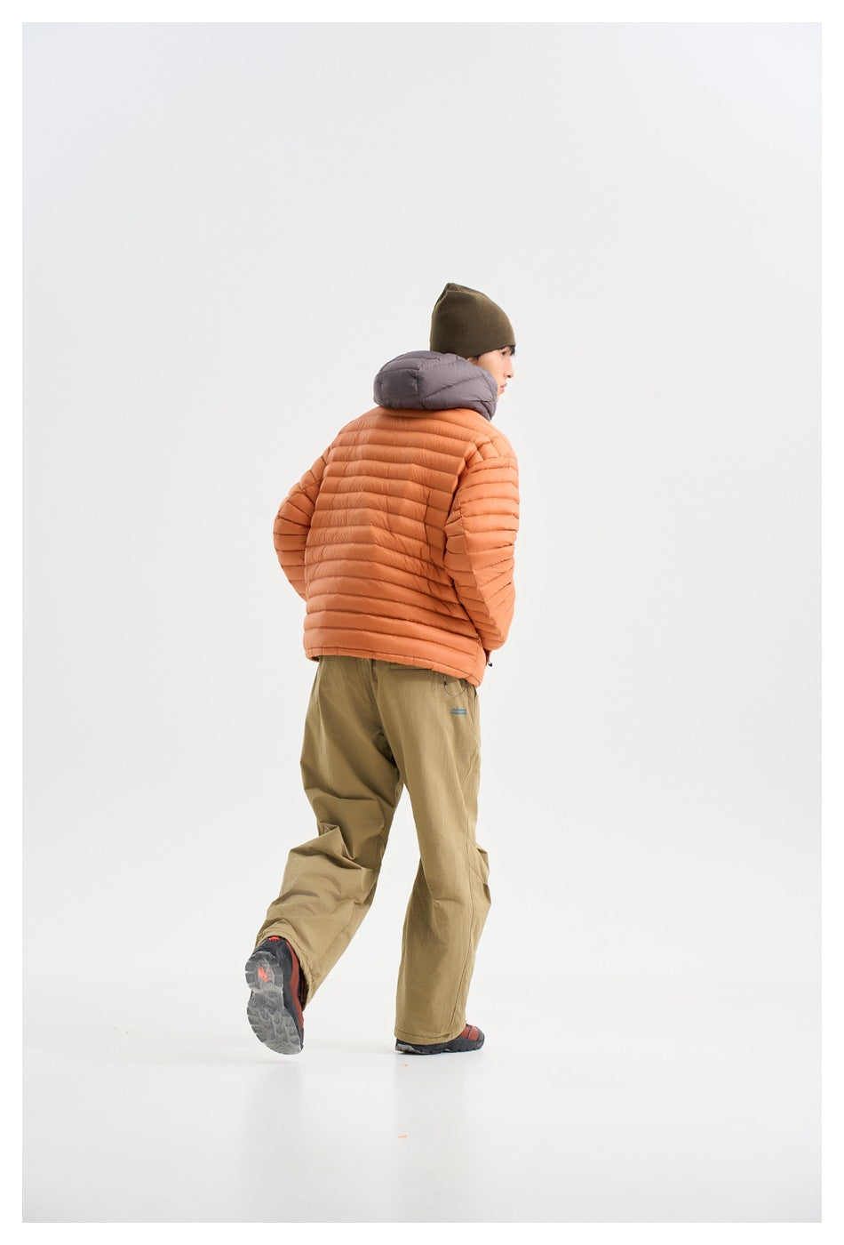 Rear three-quarter view of a model wearing an orange quilted jacket with a gray hood and loose olive trousers.