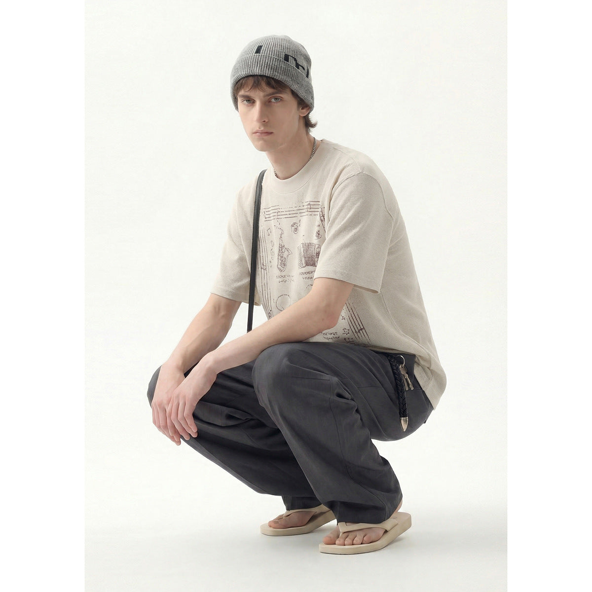 Model crouching in an ivory graphic T-shirt, charcoal trousers and a gray beanie.