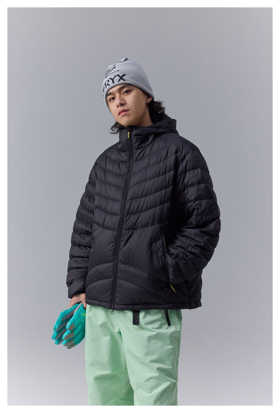 Three-quarter view of a model wearing a black hooded quilted jacket and mint-green snow pants.
