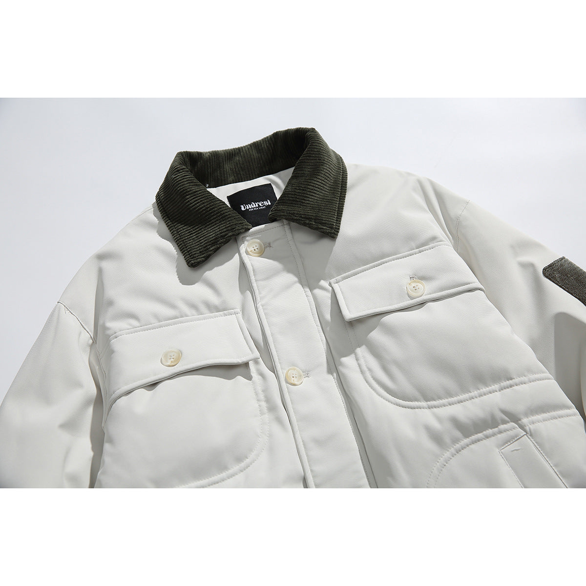 Close-up of an off-white jacket with a dark ribbed collar, buttoned flap chest pockets and a sleeve patch.