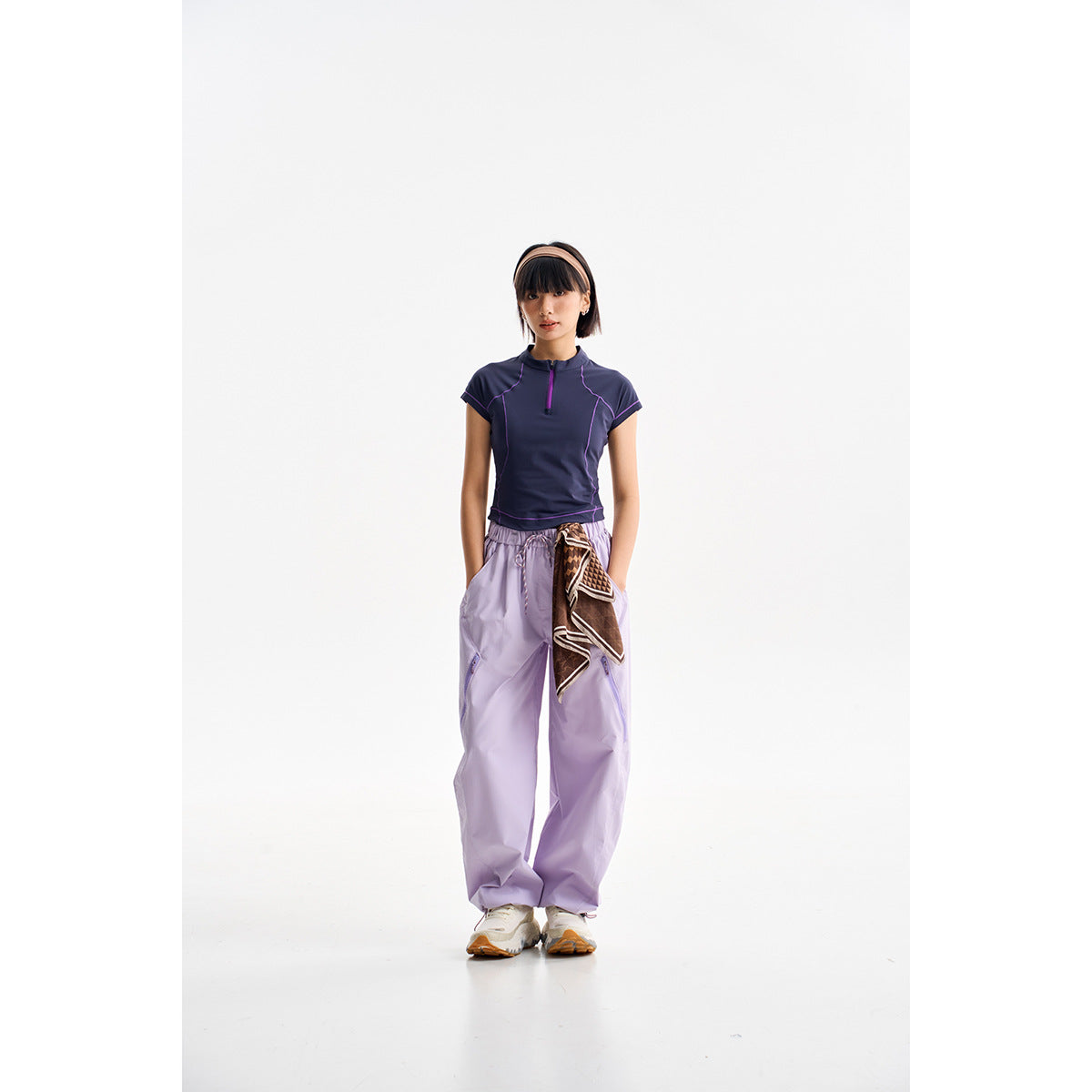 Model wearing a navy short-sleeve quarter-zip top with purple seams and loose lavender pants, with a patterned scarf at the