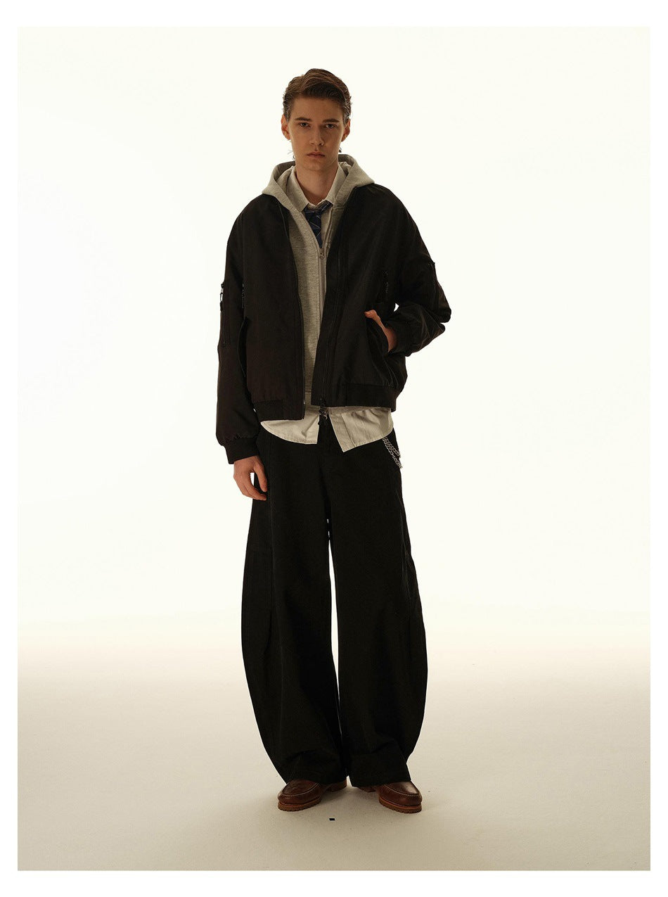 Model wearing a black zip bomber jacket over a gray hoodie and shirt, with wide black trousers.