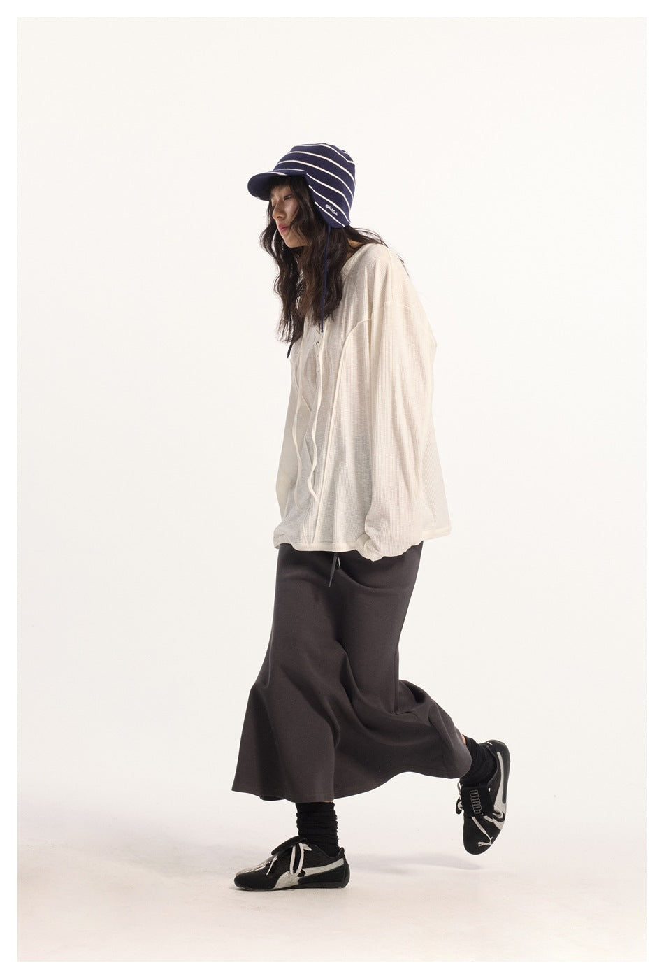 Side view of a model wearing an ivory oversized lace-up top and dark flared maxi skirt with a navy striped cap.