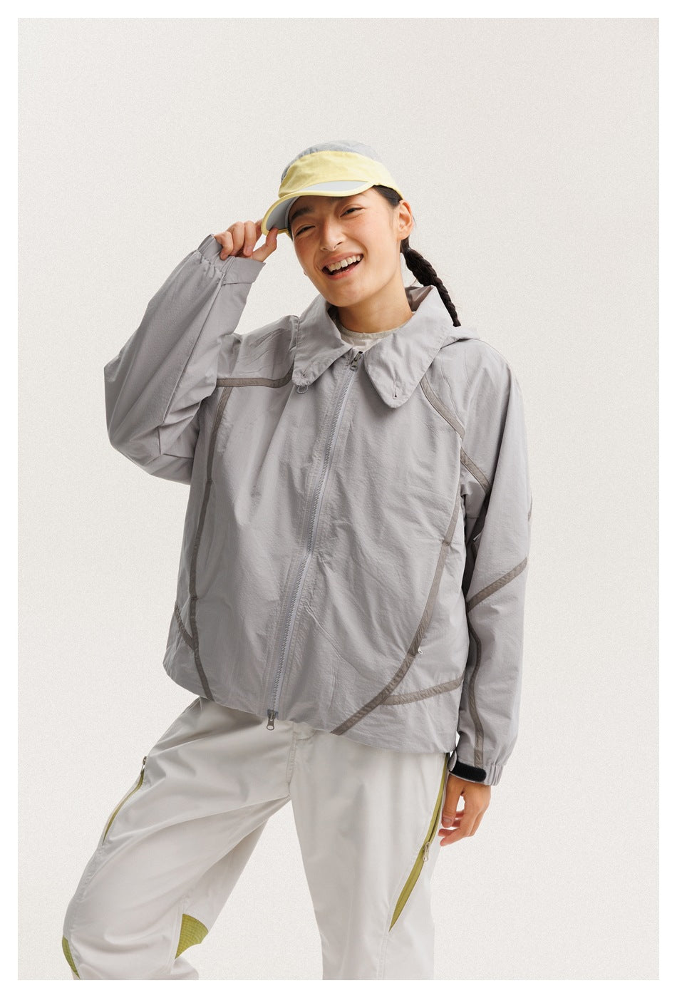 Closer view of a model wearing a light gray hooded zip jacket with dark seam details and white pants with olive accents.