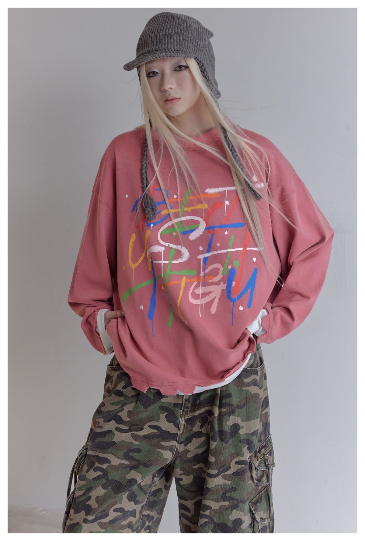 Model wearing a dusty rose oversized graphic sweatshirt with camouflage cargo pants and a gray knit cap.