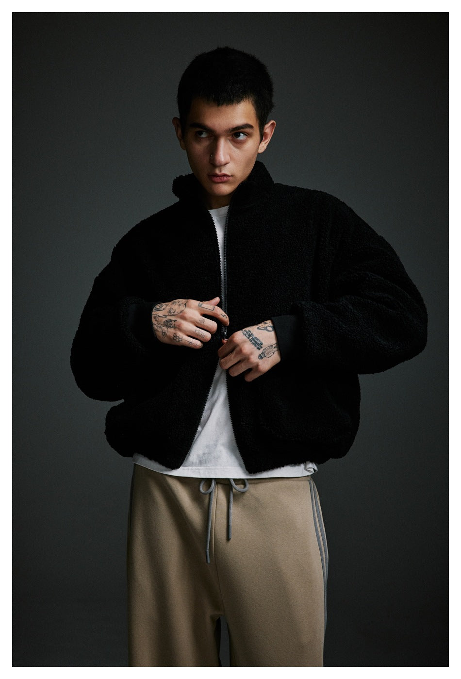 Model adjusting a black textured zip-up jacket over a white shirt with taupe drawstring pants.