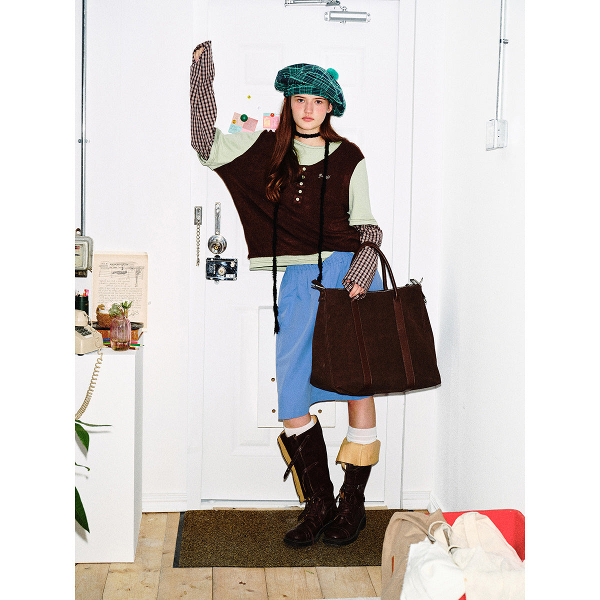 Model poses in a green plaid beret, layered sage and burgundy top with checked sleeves, blue skirt, and dark boots.