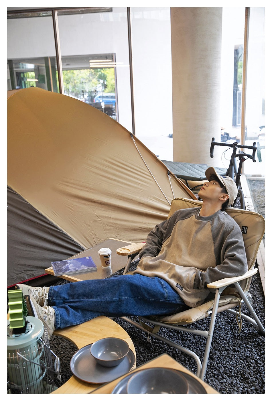 A man reclines in a camping chair wearing a taupe sweatshirt with gray raglan sleeves and blue jeans.