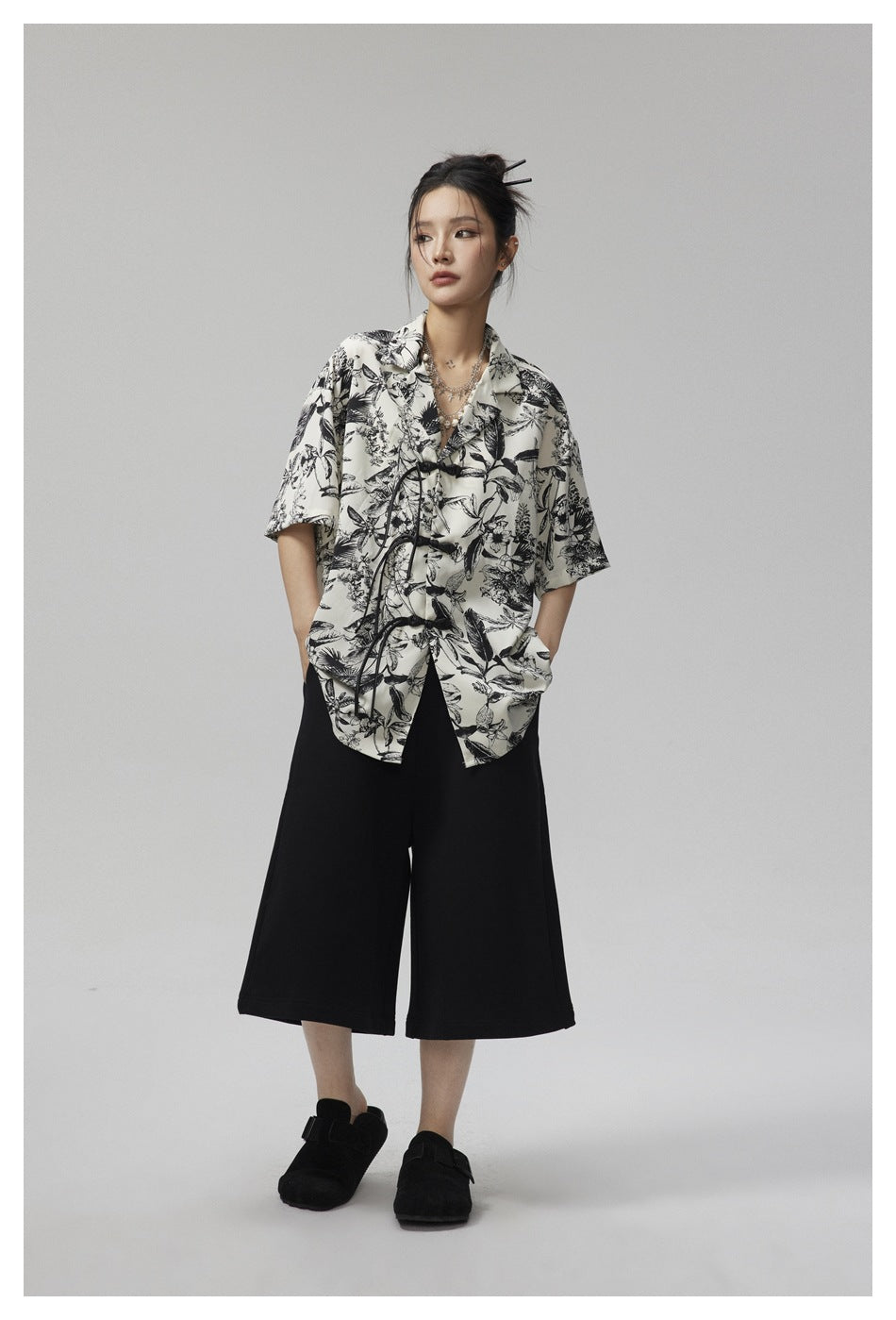 Model in the botanical print shirt with knotted frog closures, one hand in pocket, with black cropped pants