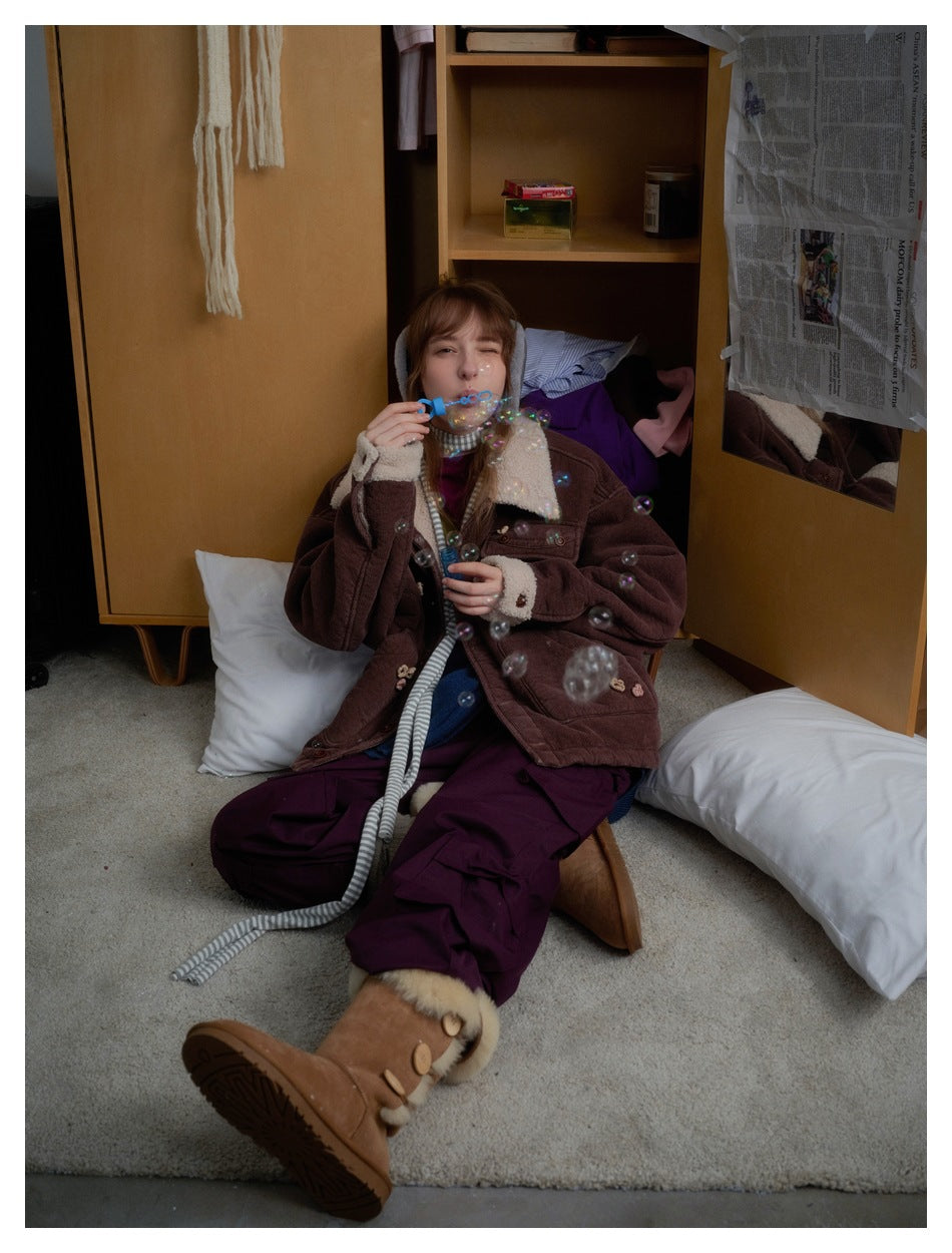 Seated model wearing a brown jacket with cream shearling trim and flower appliqués, styled with loose trousers and boots.
