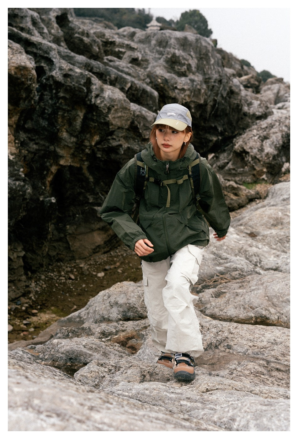 Front view of a model wearing a dark green hooded jacket and cream cargo pants on rocky terrain.
