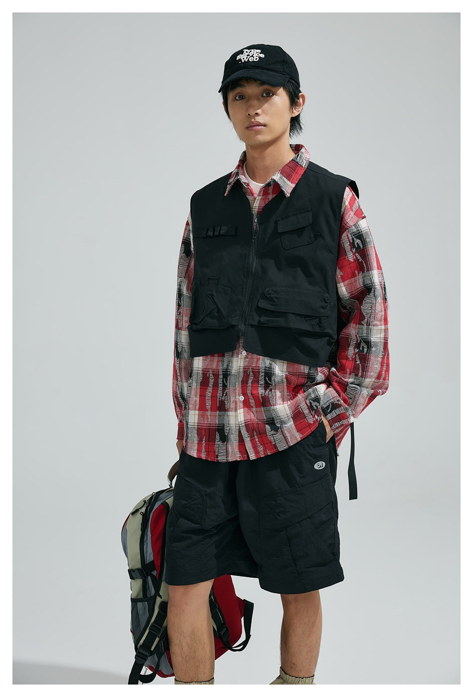 Model in a black multi-pocket utility vest over a red plaid shirt and black shorts.