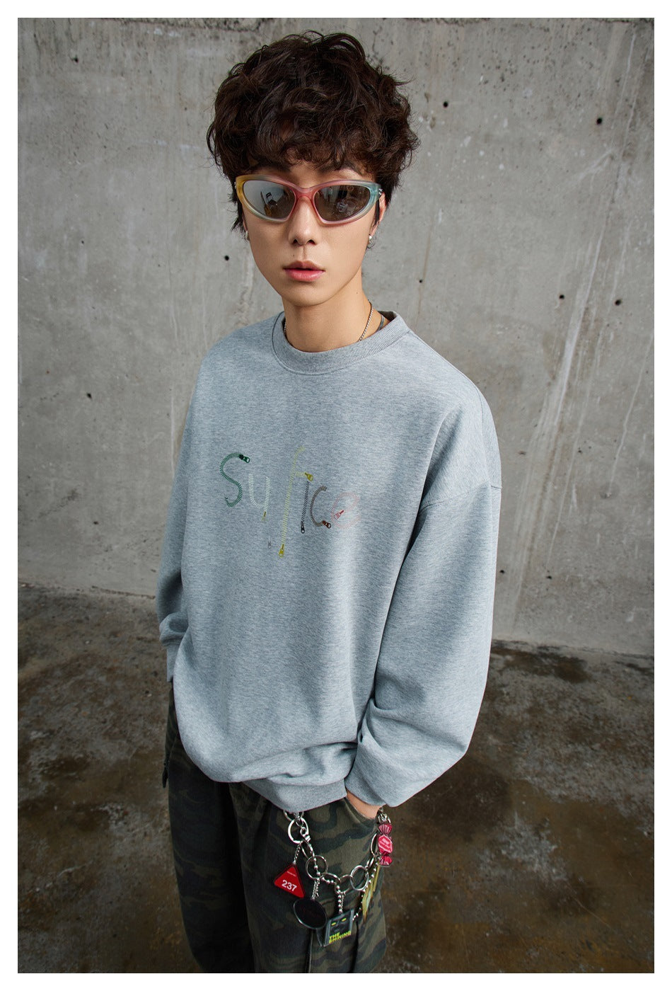 Model wearing a gray crew-neck sweatshirt with zipper-style lettering graphic, sunglasses and camouflage pants