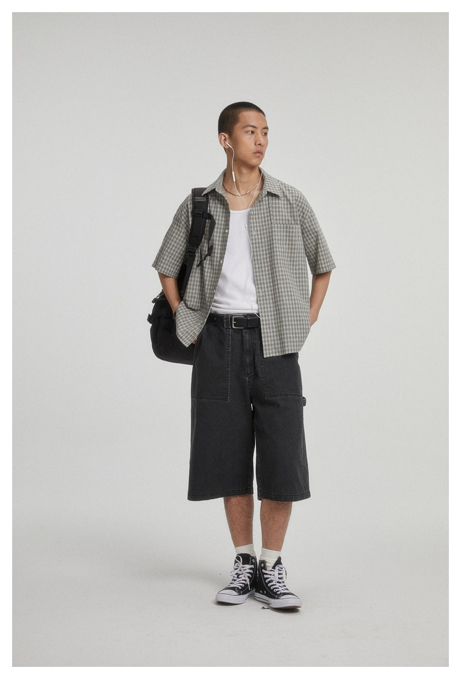 Model wearing an open gray checked short-sleeve shirt over a white tank top with dark shorts and a shoulder bag.