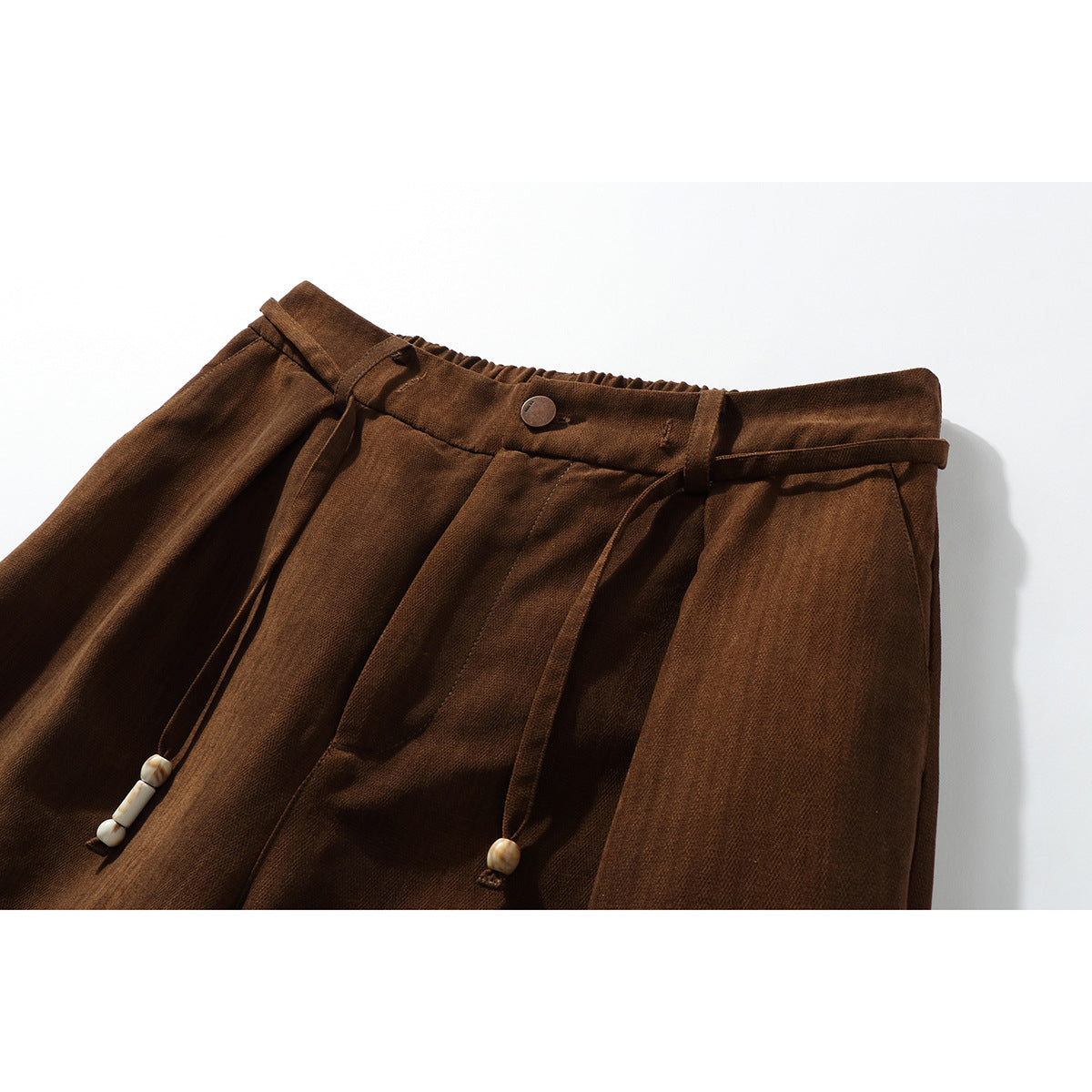 Close-up of brown trousers showing the gathered waistband, button, belt loops, pleats and dangling cords with pale beads.