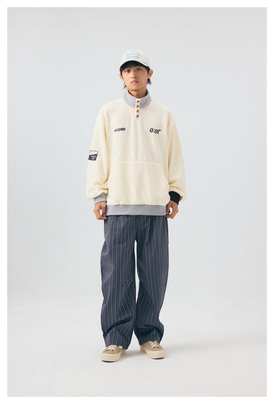 Model wearing the off-white button-collar pullover with striped wide pants, cap and tan sneakers, front view
