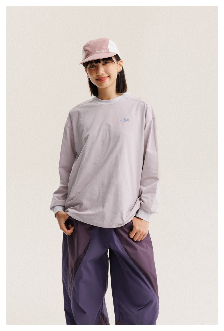 Model wearing a pale lilac long-sleeve shirt with a small chest emblem, loose purple pants, and a pink cap.