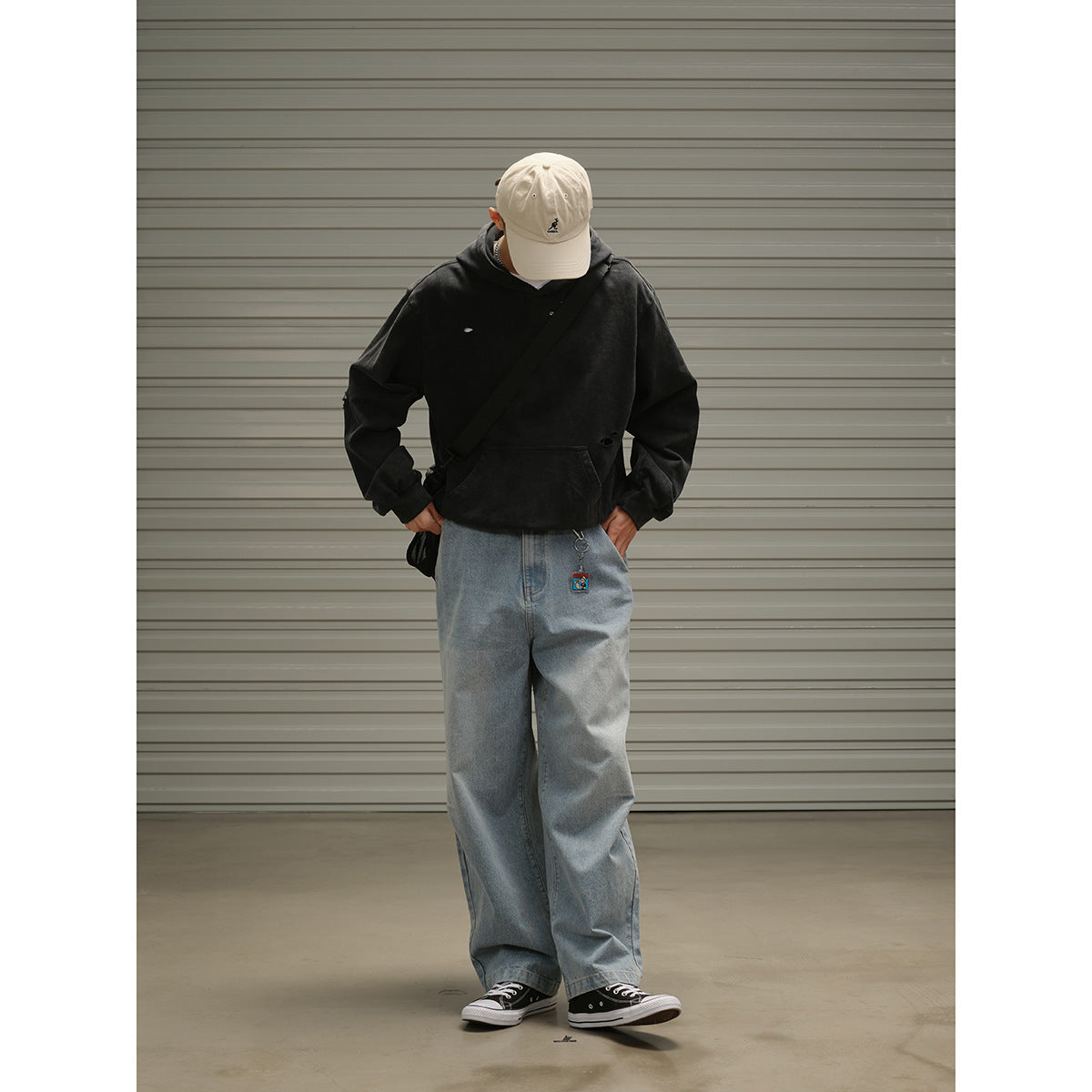 Front view of a model in a black distressed hoodie and loose blue jeans, wearing a cream cap and crossbody bag.