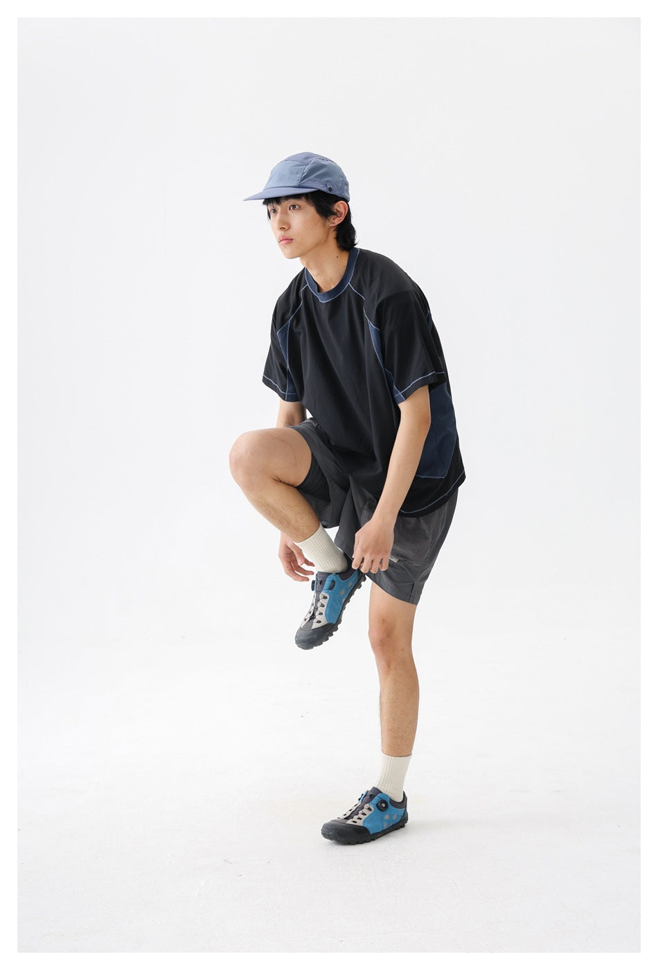 A man wearing a black and navy color-block T-shirt and gray shorts lifts one foot against a white studio background.