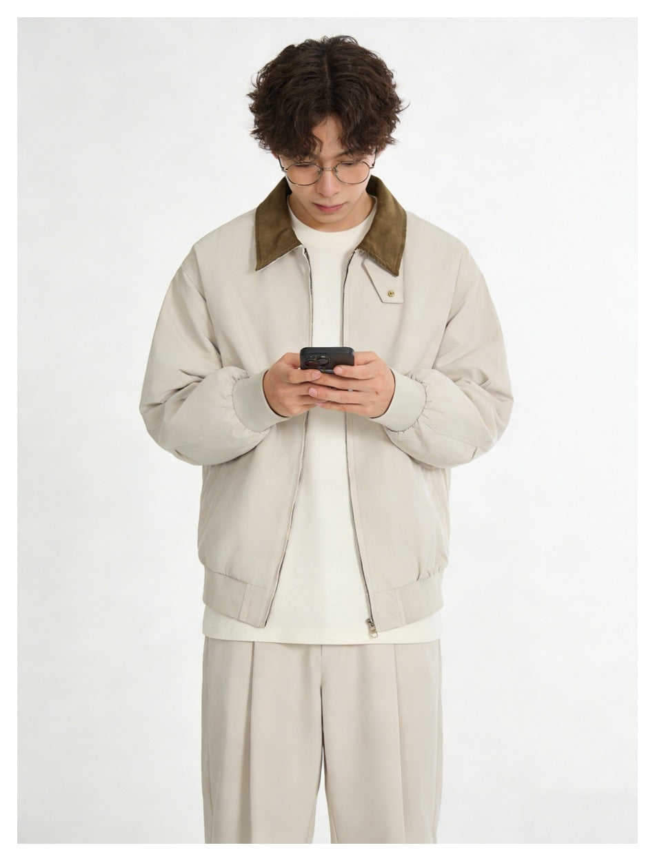 Man in beige zip jacket with brown collar over a cream tee, looking down at a phone, with beige pleated pants