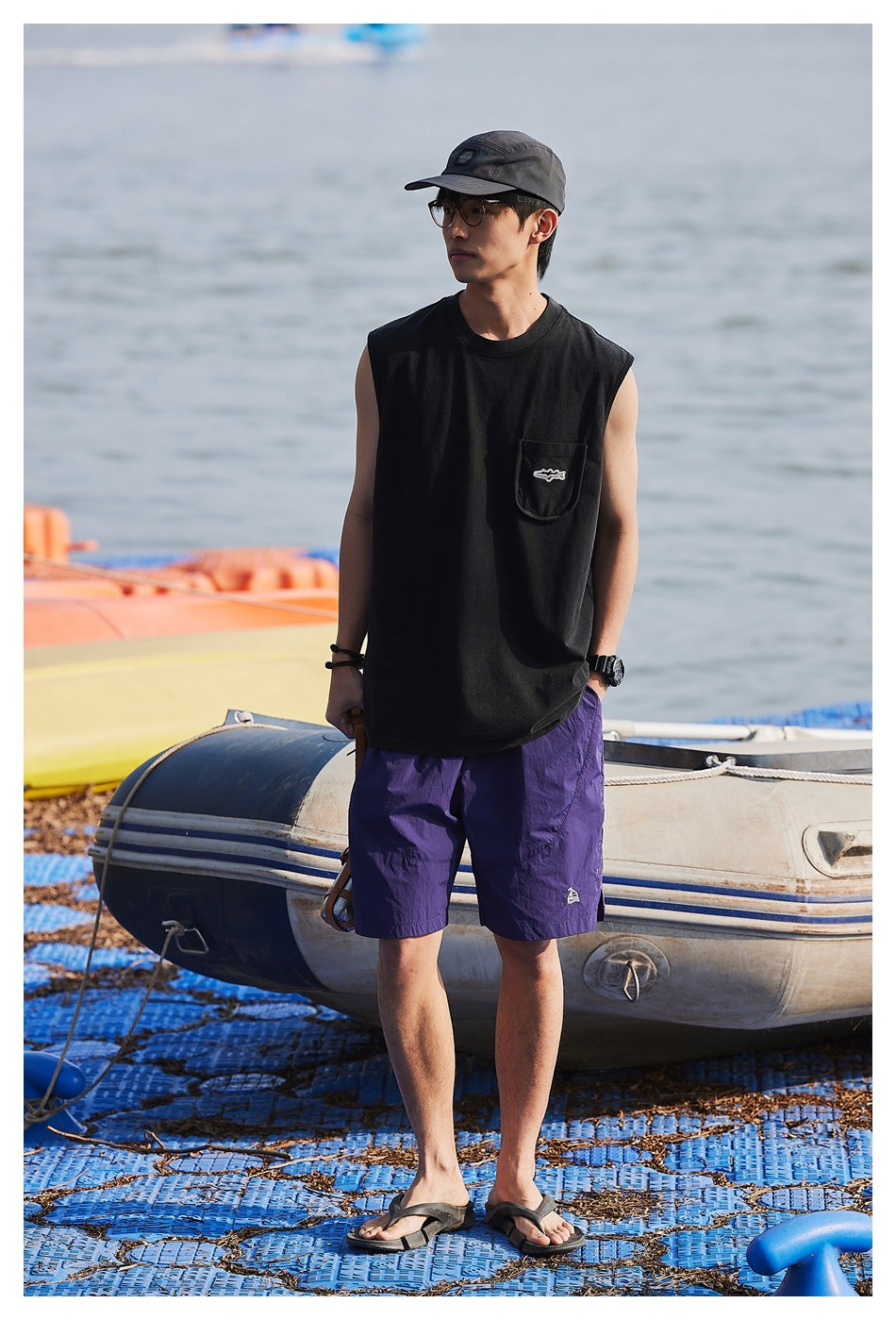 Model wearing a black sleeveless pocket shirt and purple shorts by the water, shown full length.
