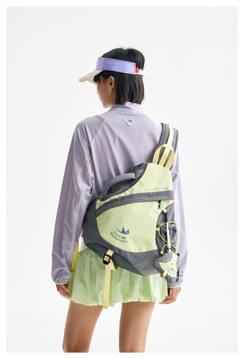 Rear view of a model in a lilac long-sleeve top and mint skirt, wearing a gray and pale yellow backpack and visor.