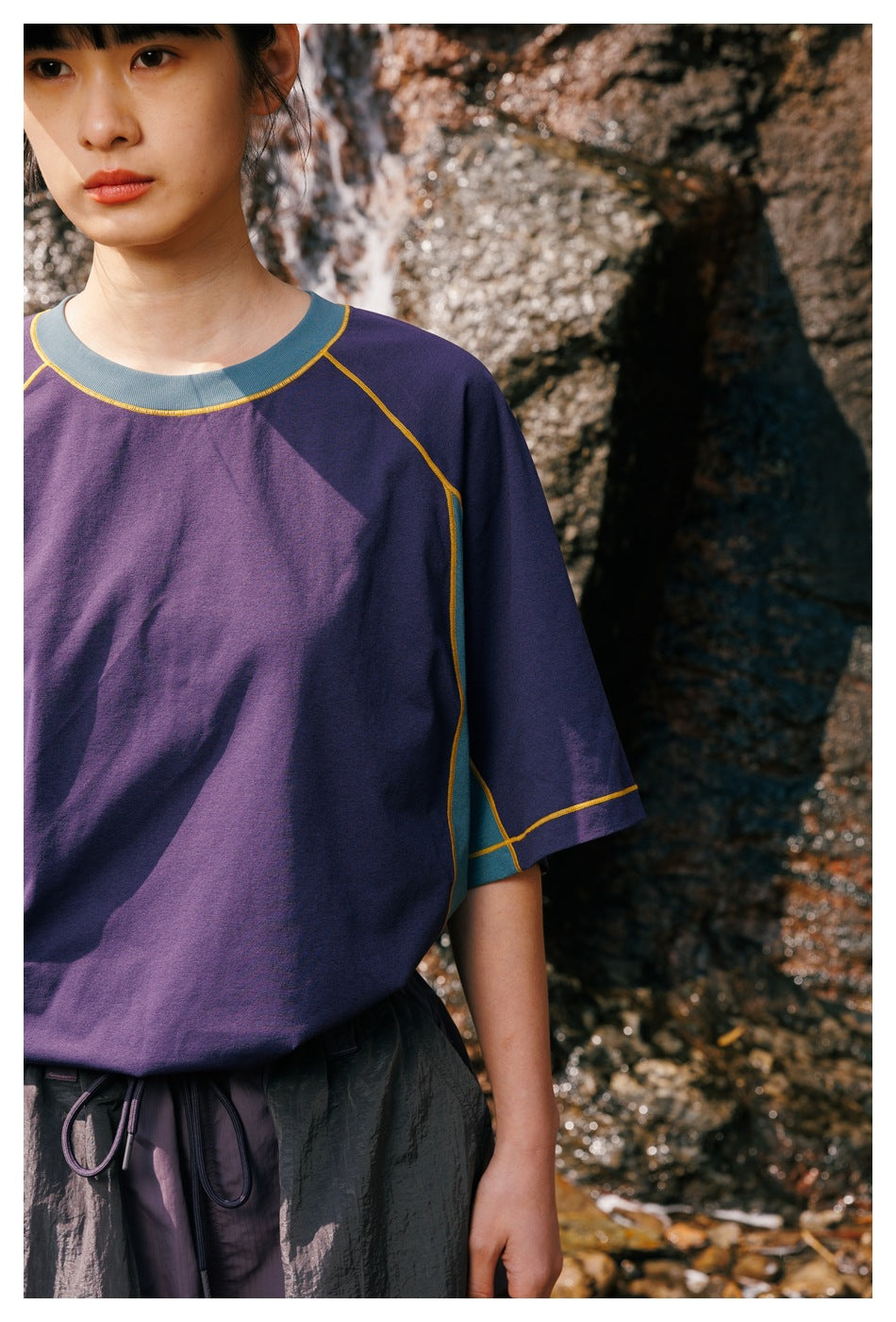 Close-up of a purple shirt showing its teal crew neck and side panels, yellow contrast stitching, and raglan sleeves.