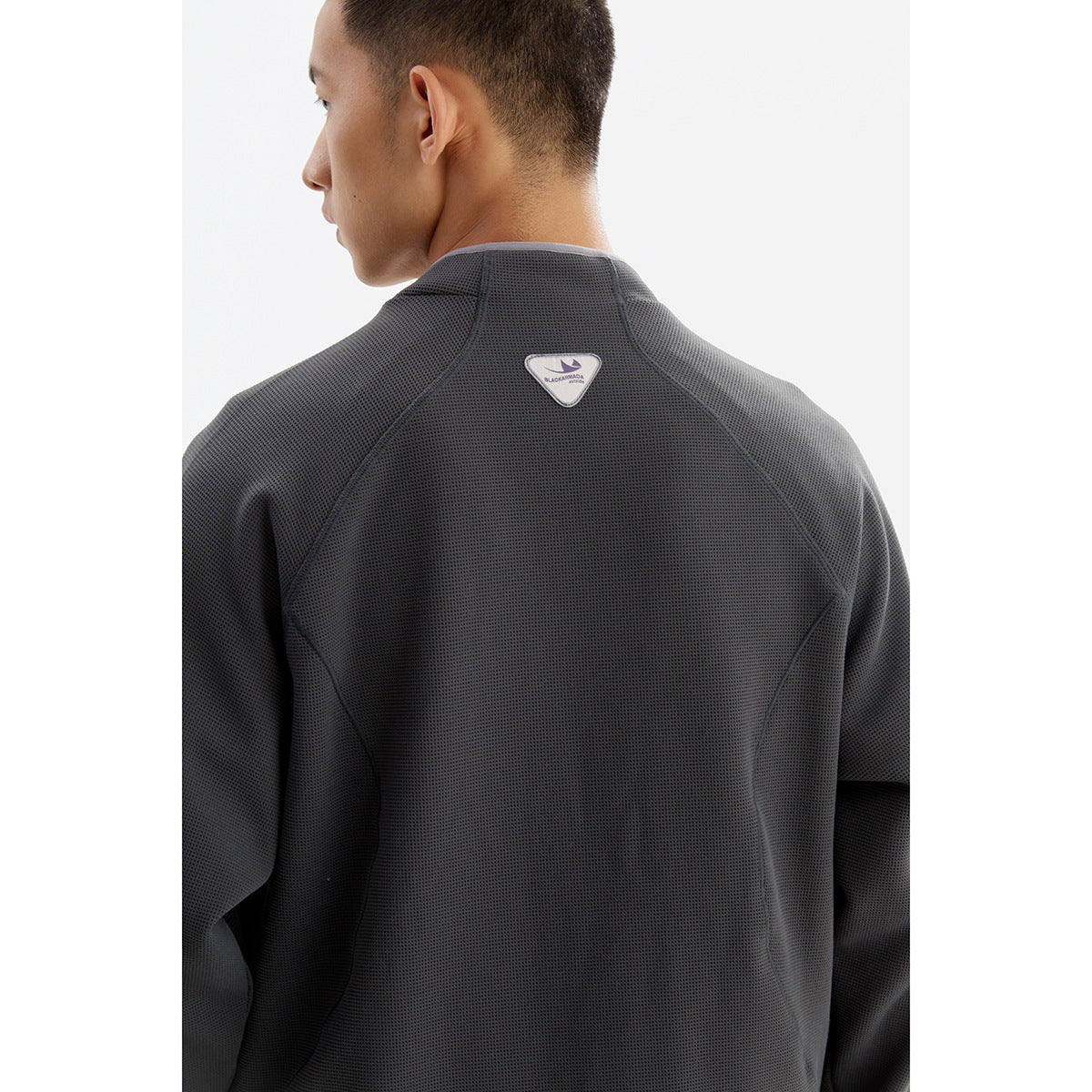 Rear view of a model wearing a charcoal textured jacket with curved seams and a small triangular upper-back patch.