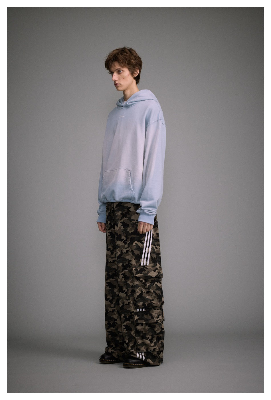 Three-quarter view of a model wearing a pale blue hoodie and loose camouflage cargo pants with white side stripes.