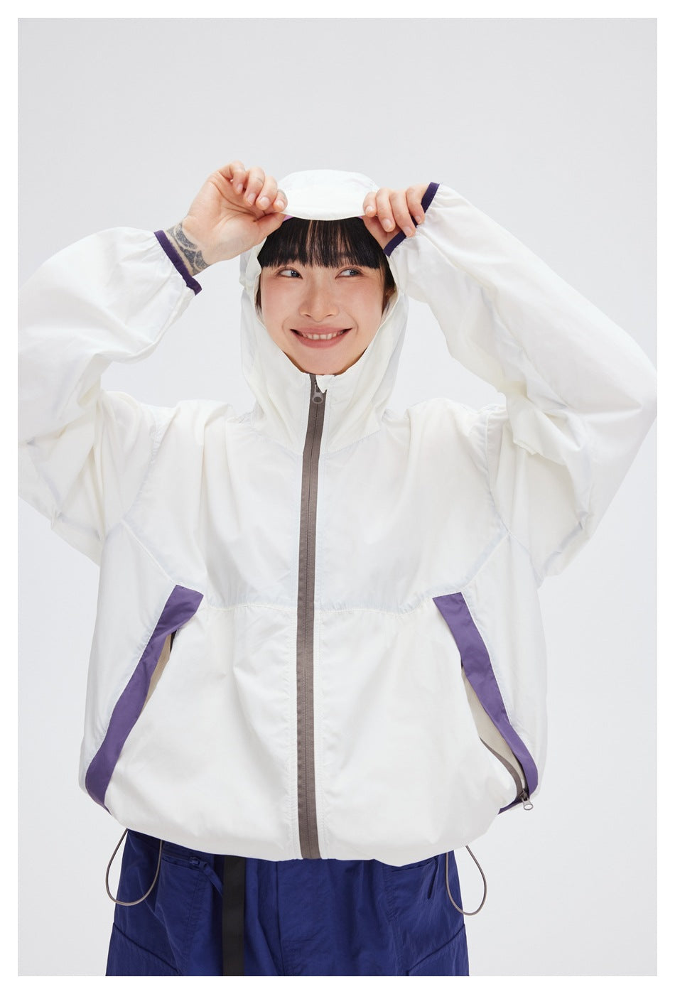 Model wearing the white hooded jacket, lifting its brim to show the purple pocket accents and gray zipper.