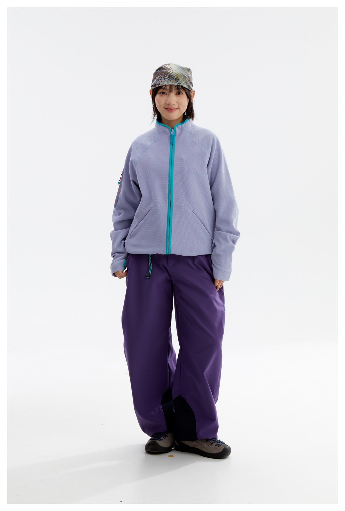 Model wearing a zipped lavender jacket with turquoise trim and loose purple pants, with a patterned cap.