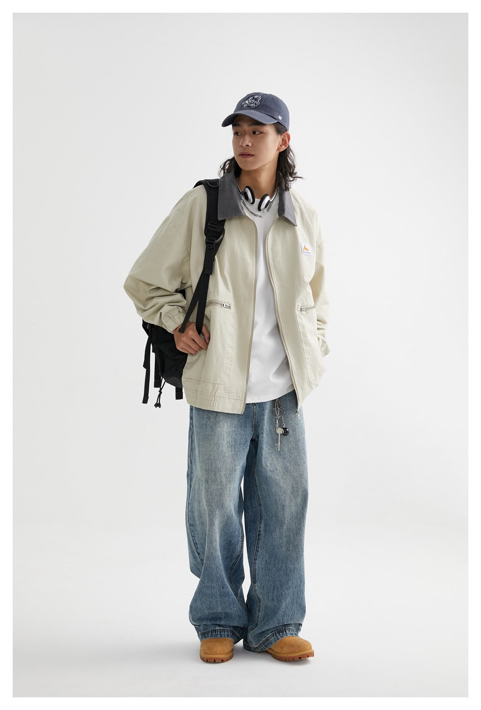 Full-length view of a model wearing a pale beige jacket with a gray collar, loose blue jeans, and a black backpack.