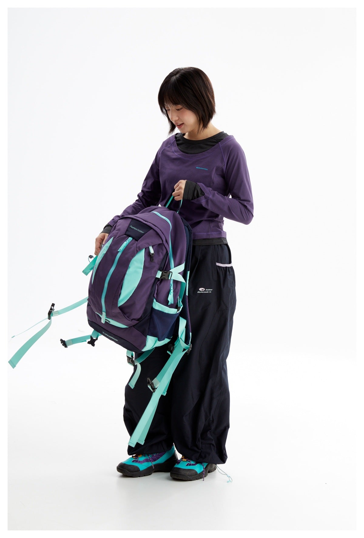 Model wearing a purple long-sleeve top and black pants while holding a purple and turquoise hiking backpack.