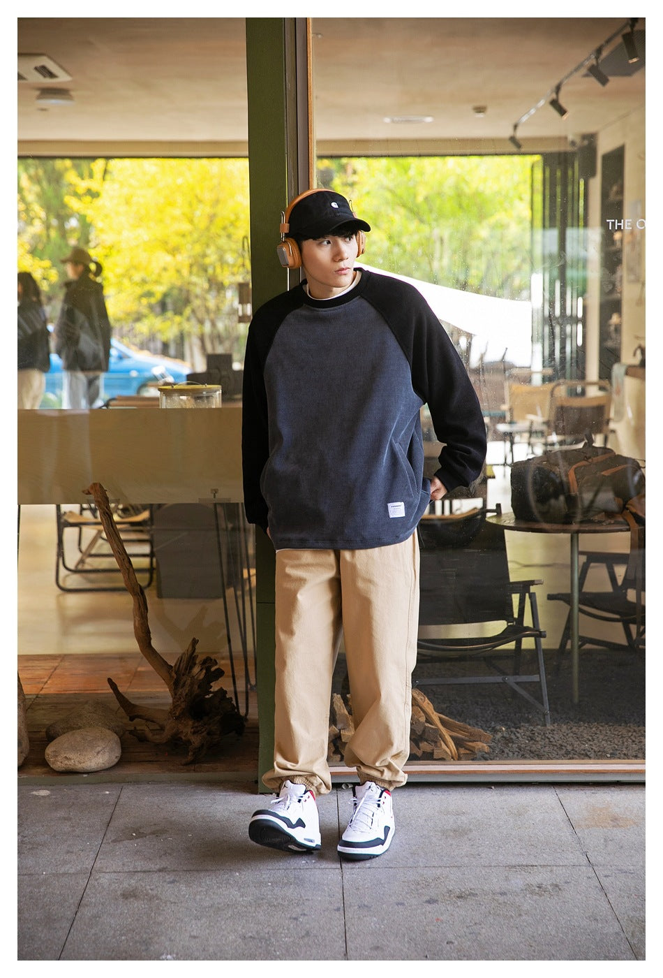 Model wearing a gray and black raglan crewneck sweatshirt with tan pants, cap, headphones and sneakers outside a cafe