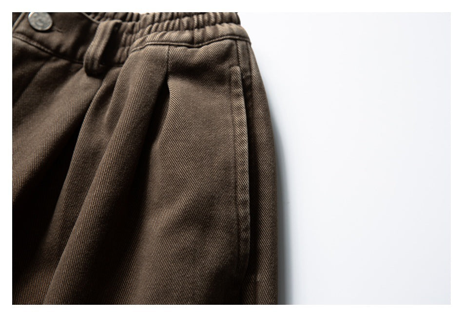 Close-up of brown trousers showing the gathered elastic waistband, button, and front pocket seam.