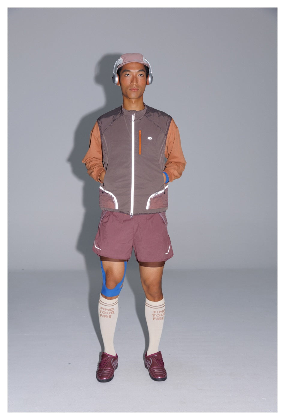 Full-length model wearing a taupe sleeveless zip vest, rust long-sleeve top, and mauve athletic shorts.