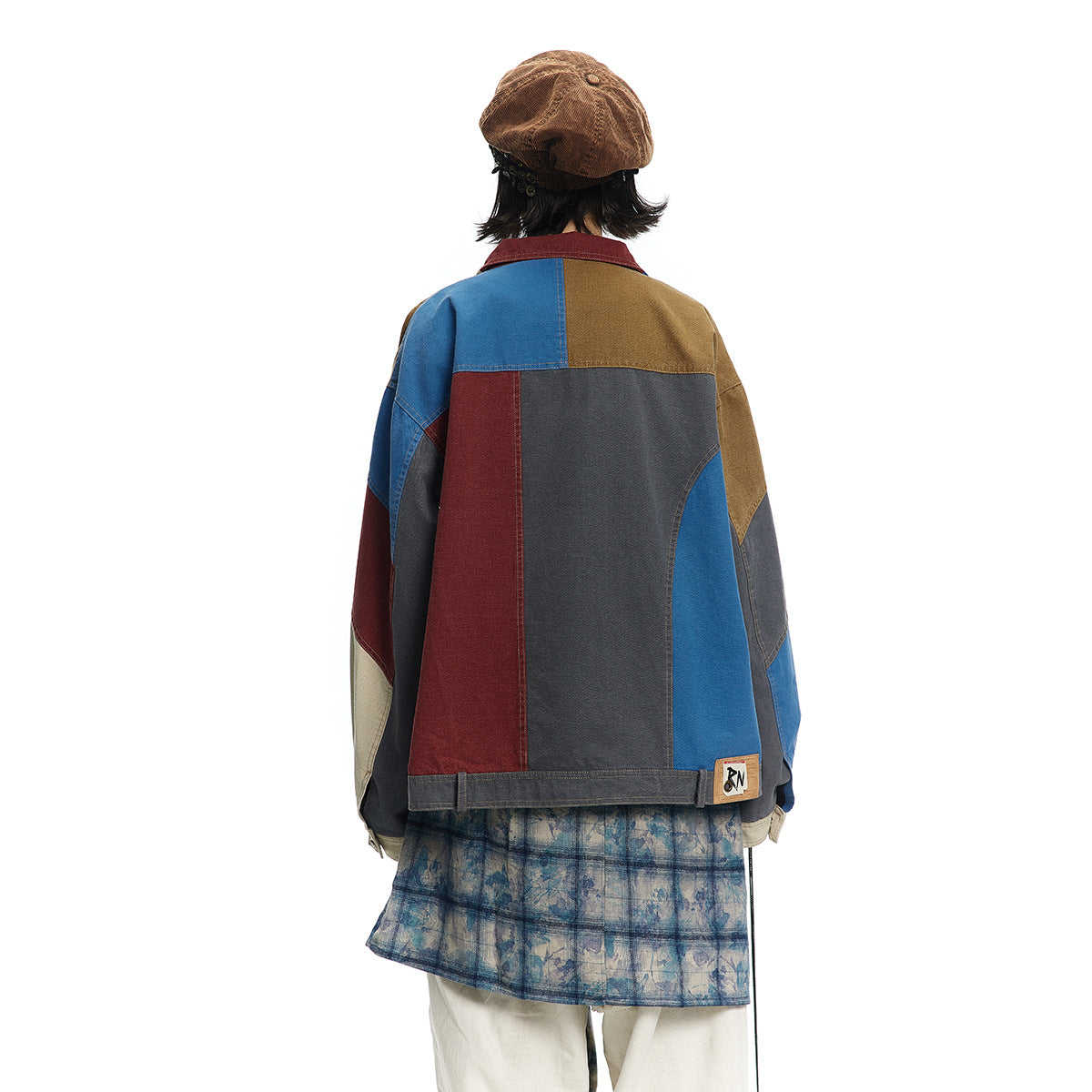 Back view of a model wearing a color-block patchwork jacket over a blue patterned skirt and cream trousers.