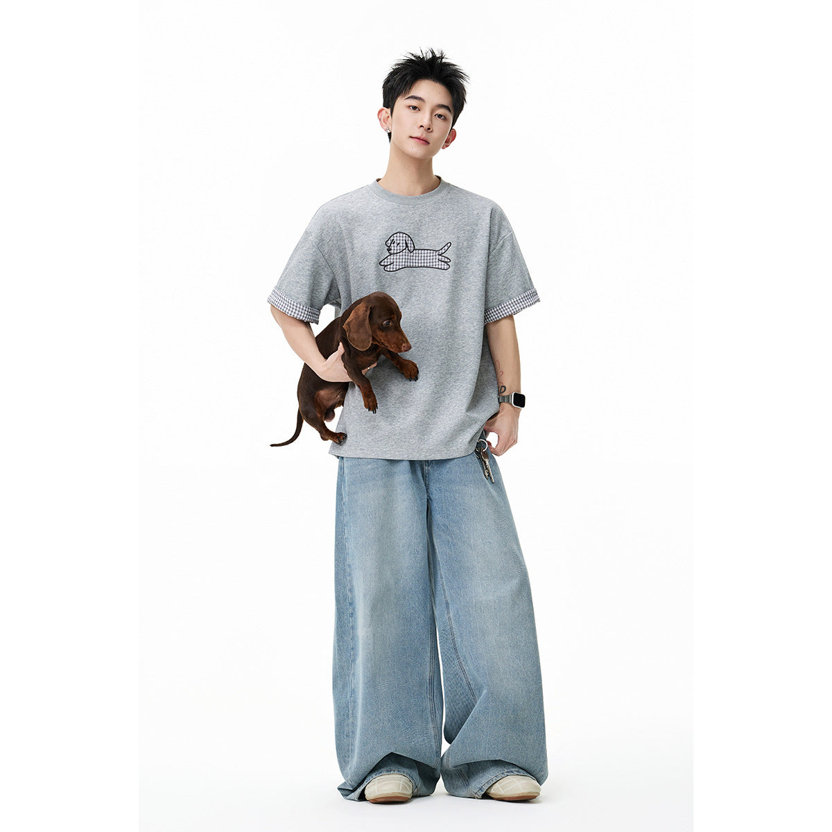 Full-length view of a man wearing a gray dog-graphic T-shirt with checked cuffs and wide-leg light blue jeans.
