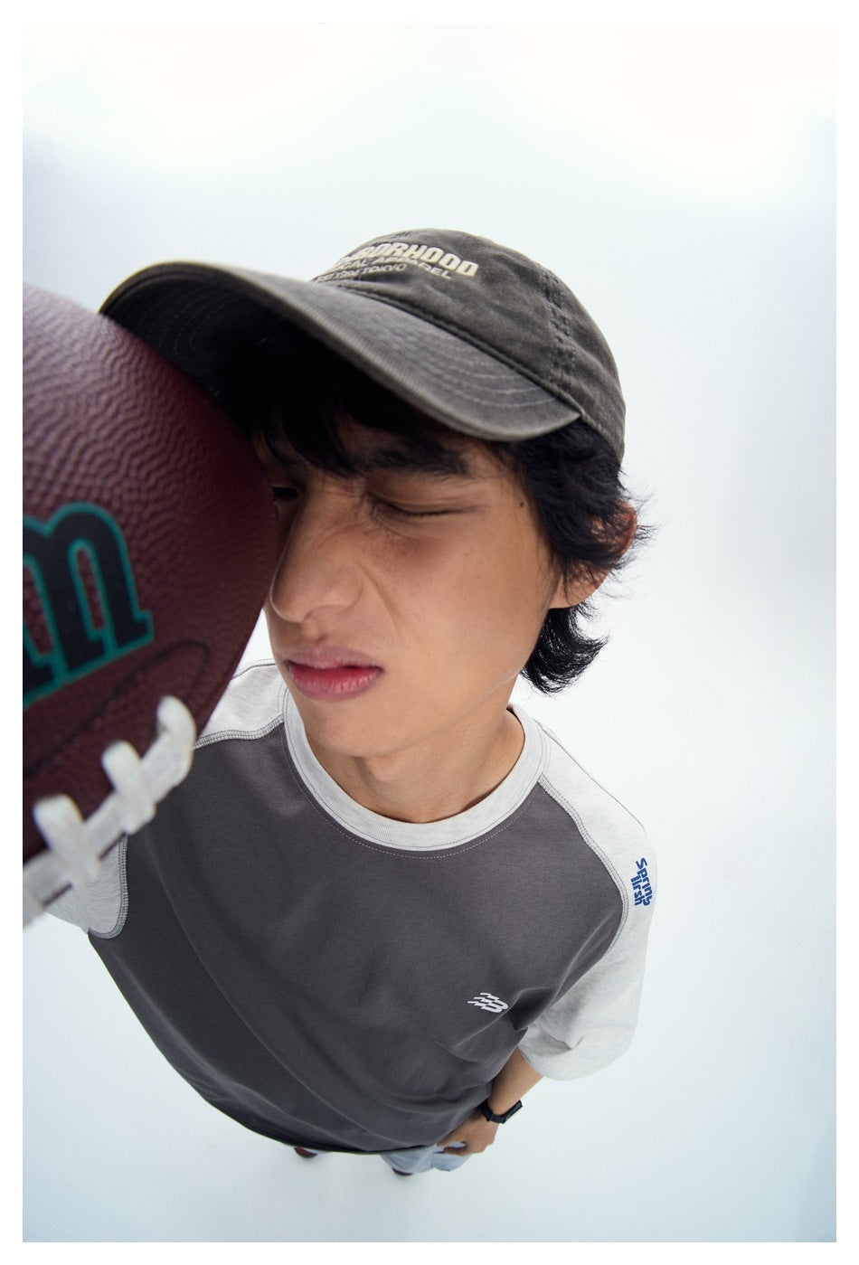 Close-up of a person in a gray raglan T-shirt with light gray sleeves, holding a football to the face