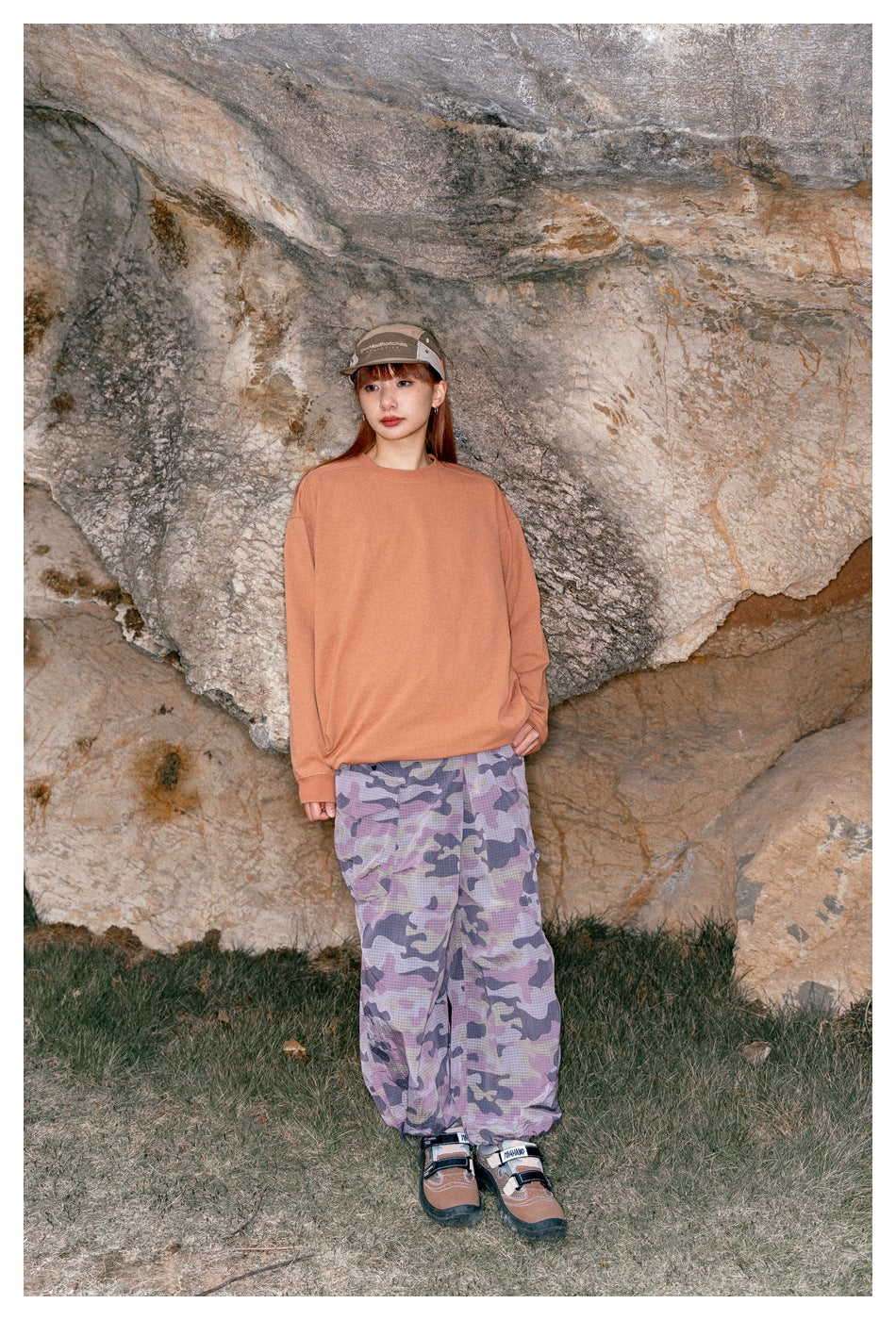 Woman standing against a rock face in a peach long-sleeve shirt and loose lavender camouflage trousers.