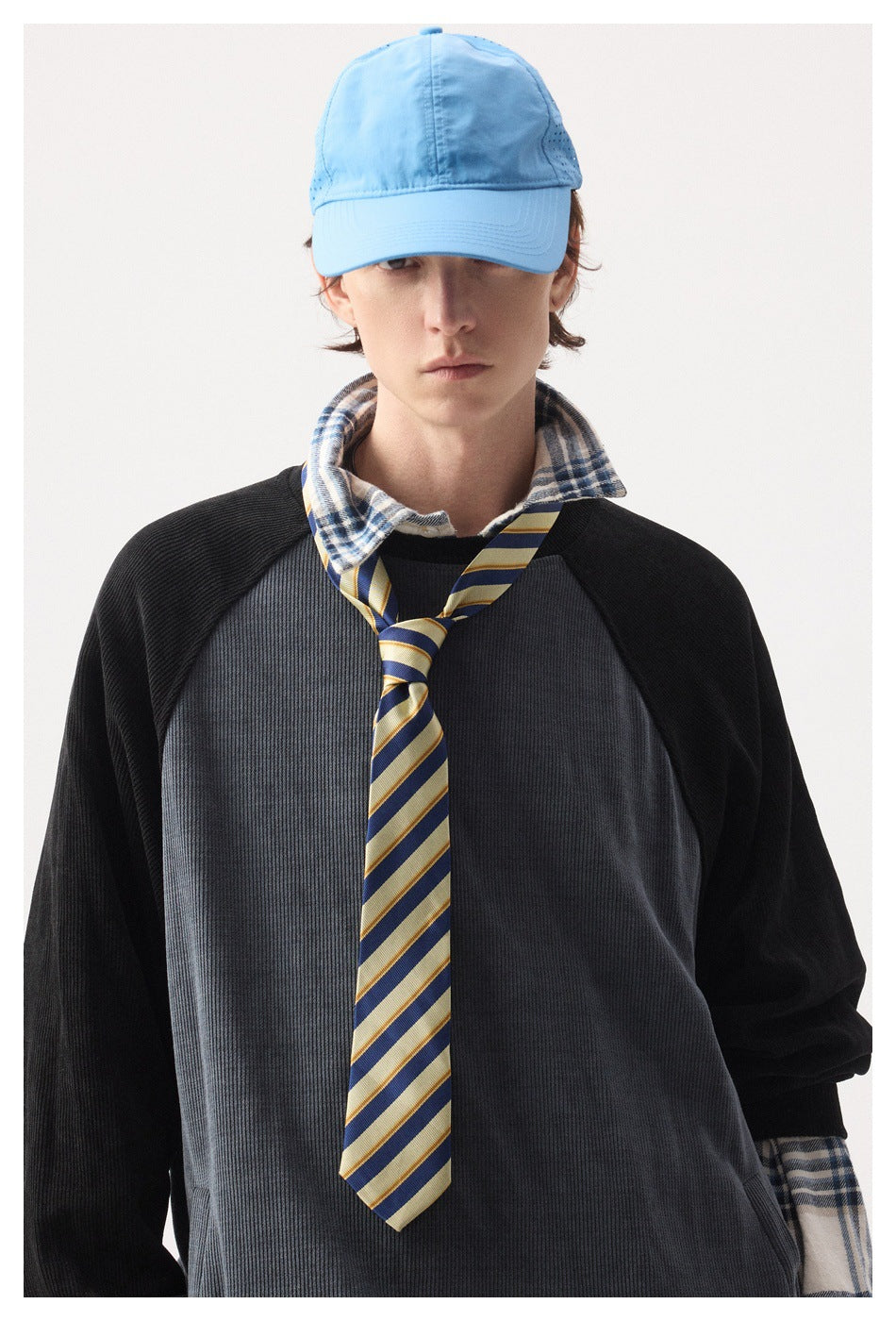Model wearing a charcoal ribbed raglan sweater over a plaid shirt, with a blue cap and striped tie.