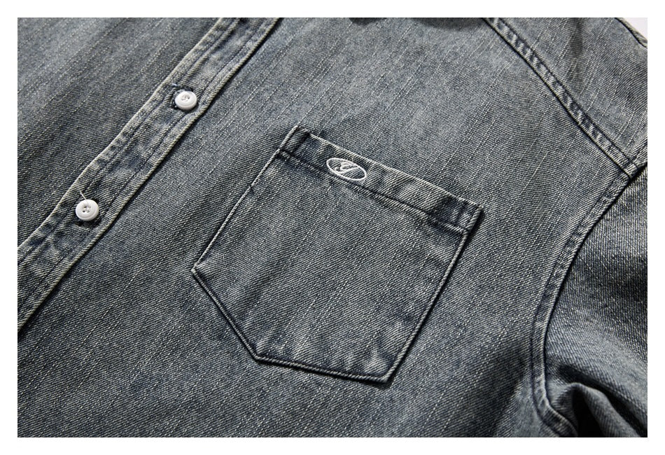 Inset close-up of a dark gray washed denim shirt chest pocket with a small embroidered oval and white buttons.