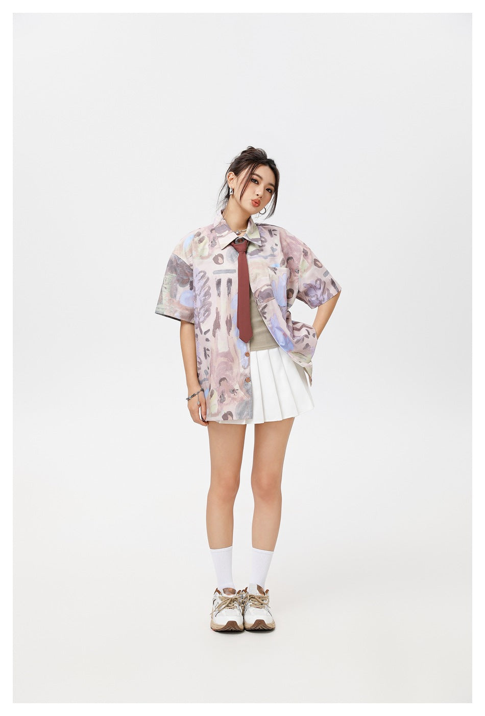 Full-length model wearing a pastel abstract-print oversized button-up shirt, burgundy tie, white pleated skirt and sneakers.