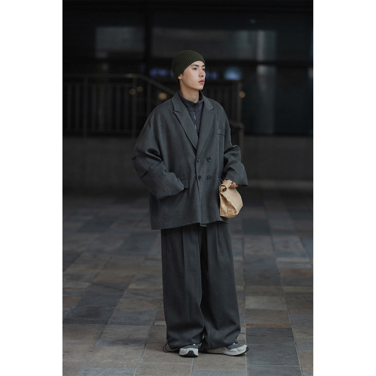Model wearing an oversized charcoal gray double-breasted coat with wide-leg trousers and an olive beanie.