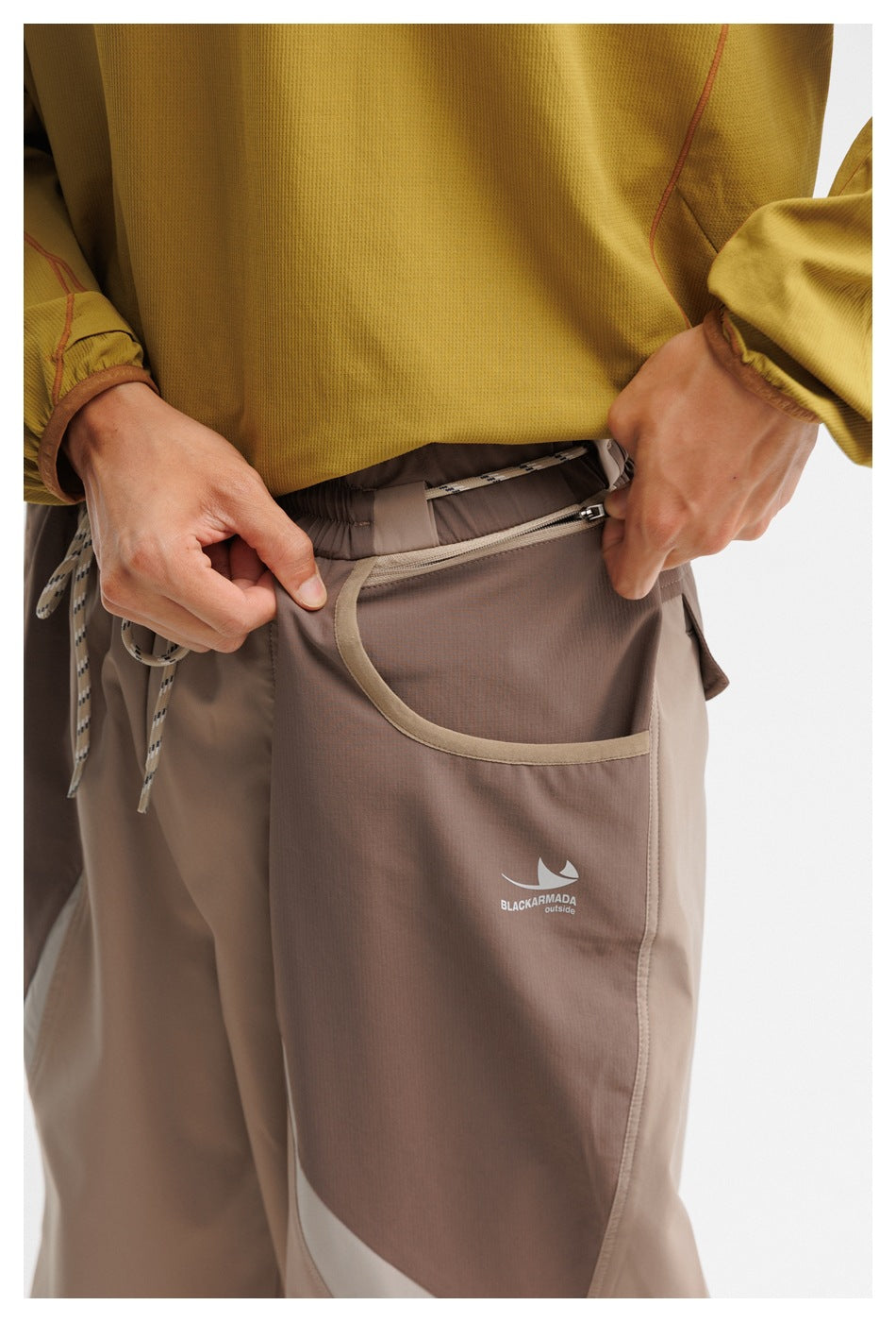 Close-up of taupe trousers showing the curved pocket, zippered opening, drawcord waist, and small garment logo.