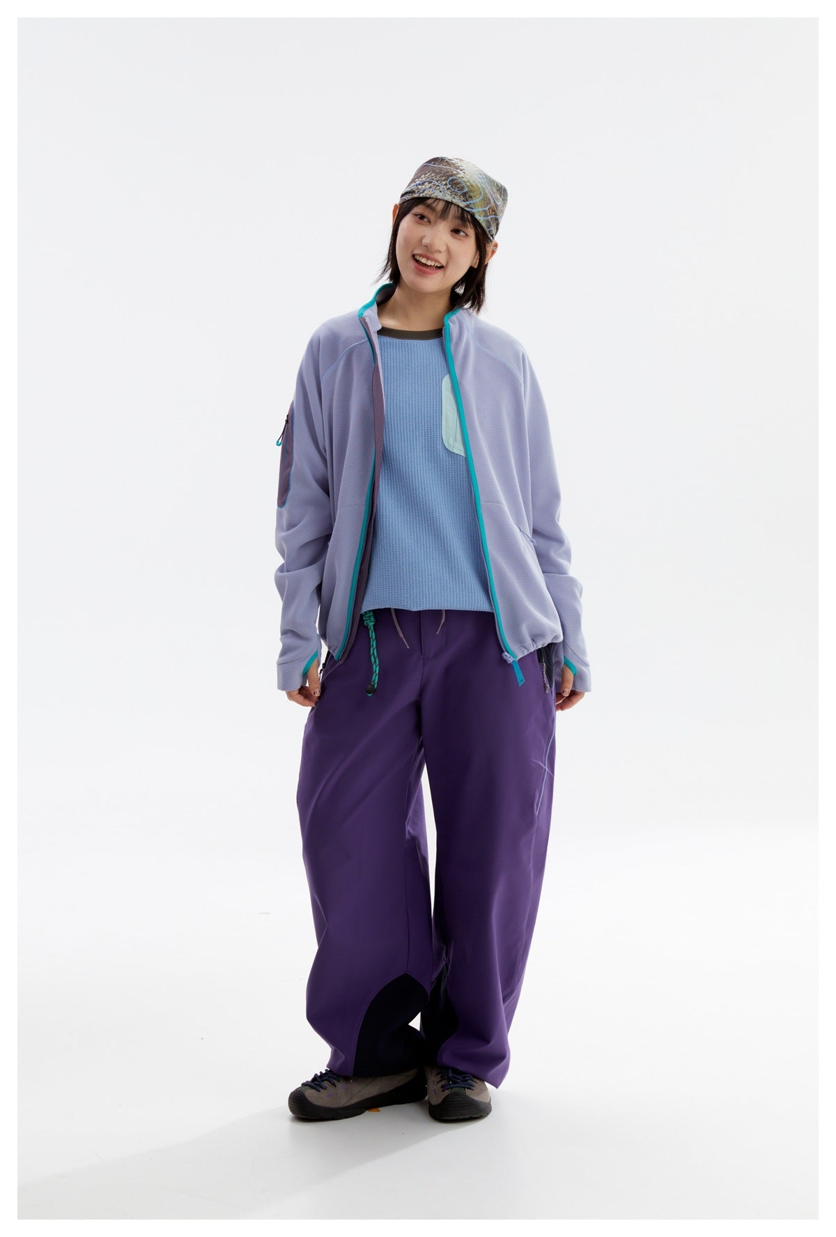 Model wearing an open lavender jacket with turquoise trim, a blue top, loose purple pants and a patterned cap.