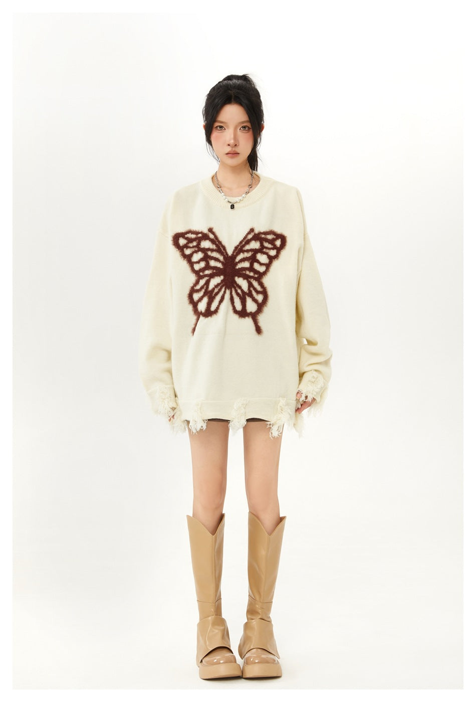 Model wearing an oversized ivory sweater with a fuzzy brown butterfly motif and frayed edges, styled with tan boots.