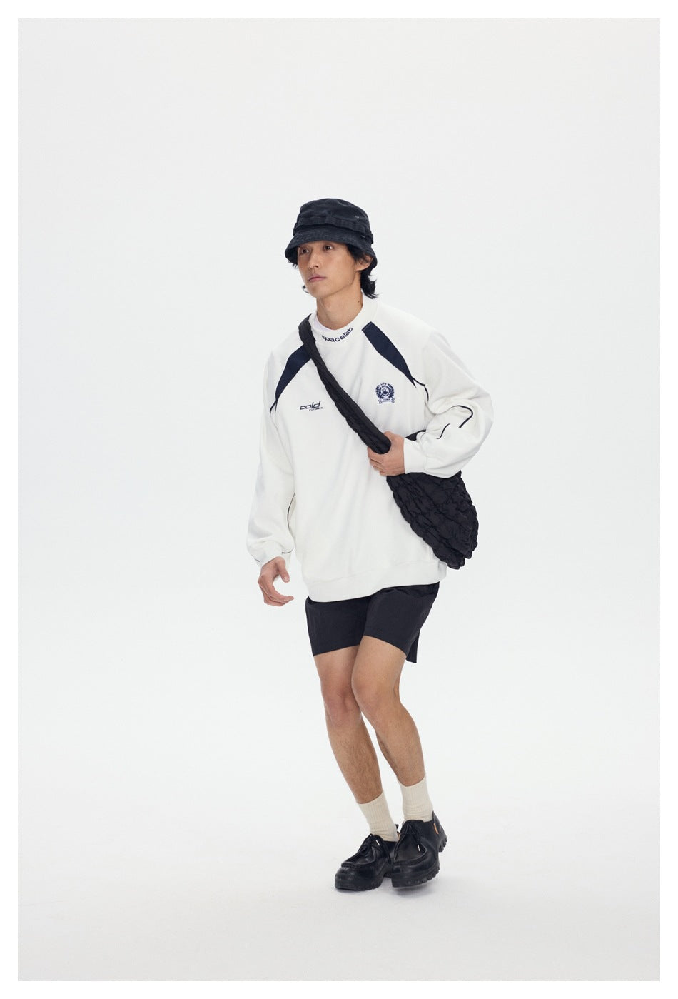 Model wearing a white graphic sweatshirt, black shorts, bucket hat and crossbody bag, shown full length.